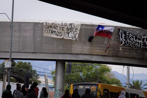 A group of people is gathered under a footbridge, where banners with political messages are displayed. The Chilean flag is visible, indicating a protest or rally. A yellow bus is also present, with visible signage.