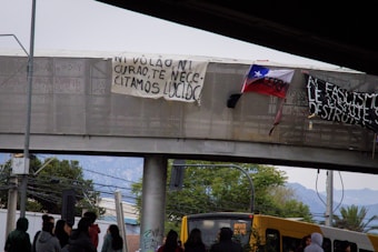 A group of people is gathered under a footbridge, where banners with political messages are displayed. The Chilean flag is visible, indicating a protest or rally. A yellow bus is also present, with visible signage.