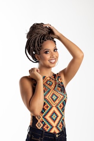 A woman with braided hair styled into an updo smiles while holding her hair. She is wearing a colorful, geometric-patterned top and dark jeans, standing against a plain, light background.