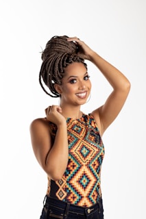 A woman with braided hair styled into an updo smiles while holding her hair. She is wearing a colorful, geometric-patterned top and dark jeans, standing against a plain, light background.