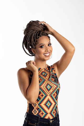 A woman with braided hair styled into an updo smiles while holding her hair. She is wearing a colorful, geometric-patterned top and dark jeans, standing against a plain, light background.