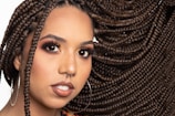A close-up portrait of a person with elaborately styled braided hair that flows to the side. They have a nose piercing, large hoop earrings, and wear makeup emphasizing their eyes and lips. The background is plain, highlighting their features.