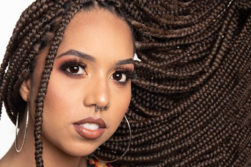 A close-up portrait of a person with elaborately styled braided hair that flows to the side. They have a nose piercing, large hoop earrings, and wear makeup emphasizing their eyes and lips. The background is plain, highlighting their features.