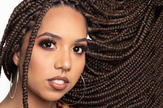 A close-up portrait of a person with elaborately styled braided hair that flows to the side. They have a nose piercing, large hoop earrings, and wear makeup emphasizing their eyes and lips. The background is plain, highlighting their features.