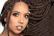 A close-up portrait of a person with elaborately styled braided hair that flows to the side. They have a nose piercing, large hoop earrings, and wear makeup emphasizing their eyes and lips. The background is plain, highlighting their features.
