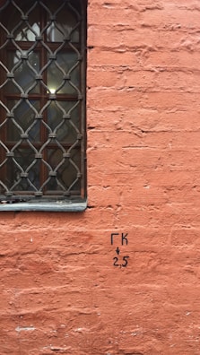 A textured orange brick wall features a window with an ornate metal grille on the left side. Black lettering reading 'ГК 2,5' with a downward arrow is visible on the brickwork.