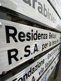 Various directional signs with black lettering on a reflective surface are mounted on a pole. The signs point towards different locations in a locale that appears to be Italian, with text indicating places like a residence and a foundation. The background shows a stone wall.