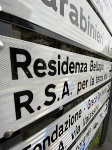 Various directional signs with black lettering on a reflective surface are mounted on a pole. The signs point towards different locations in a locale that appears to be Italian, with text indicating places like a residence and a foundation. The background shows a stone wall.