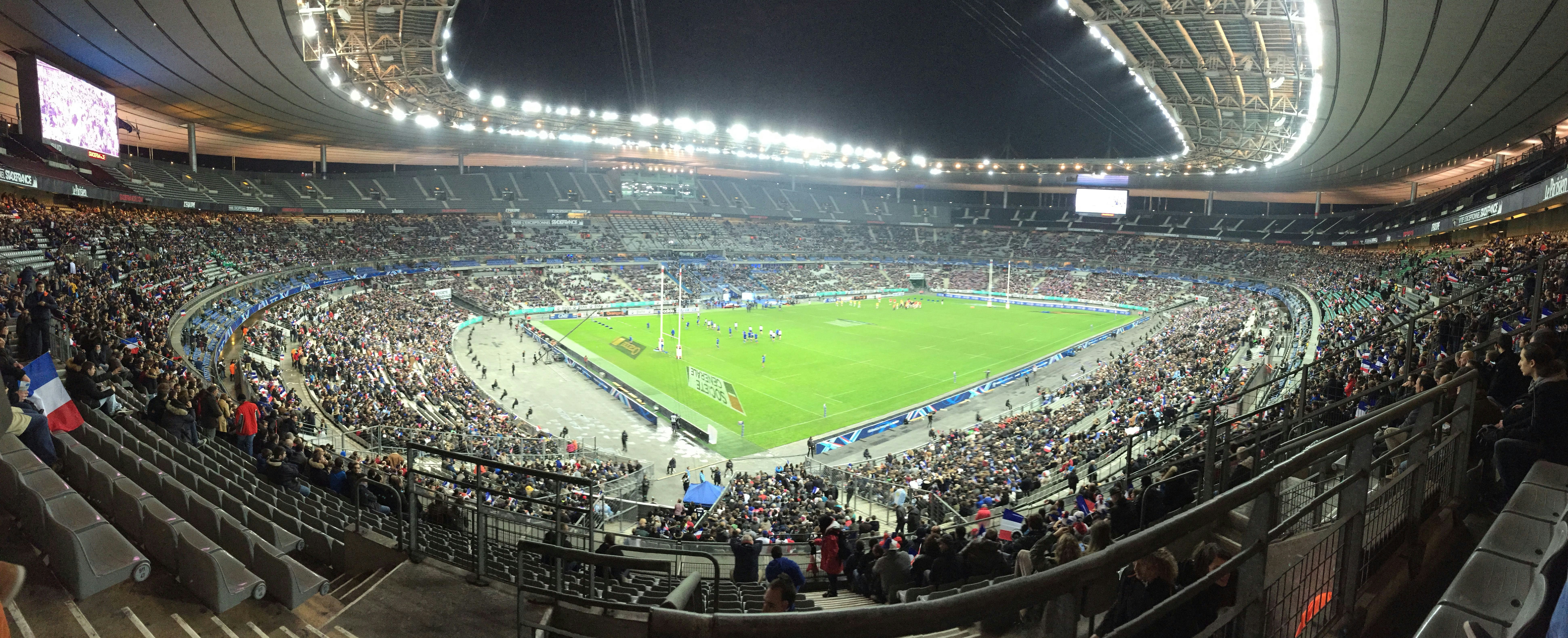 Rugby stadium packed with fans under bright stadium lighting, field visible, energetic crowd atmosphere