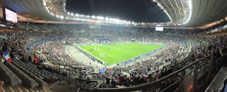 A large stadium filled with spectators, surrounding a green field where a sporting event is taking place. Bright stadium lights illuminate the area, with multiple banners and flags visible among the crowd. The architecture of the stadium is prominent, with tiered seating and large screens for viewing.