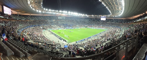 A large stadium filled with spectators, surrounding a green field where a sporting event is taking place. Bright stadium lights illuminate the area, with multiple banners and flags visible among the crowd. The architecture of the stadium is prominent, with tiered seating and large screens for viewing.