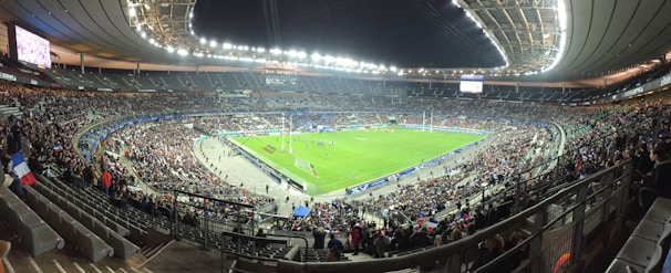 A large stadium filled with spectators, surrounding a green field where a sporting event is taking place. Bright stadium lights illuminate the area, with multiple banners and flags visible among the crowd. The architecture of the stadium is prominent, with tiered seating and large screens for viewing.