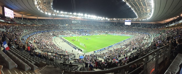A large stadium filled with spectators, surrounding a green field where a sporting event is taking place. Bright stadium lights illuminate the area, with multiple banners and flags visible among the crowd. The architecture of the stadium is prominent, with tiered seating and large screens for viewing.