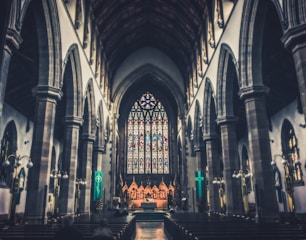 An expansive, ornate church interior featuring soaring arched ceilings and intricate stained glass windows at the far end. The dim lighting casts a serene and reverent atmosphere across the wooden pews, which are mostly empty. Ornate columns line the walkways, and a few people sit in quiet contemplation.