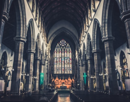 An expansive, ornate church interior featuring soaring arched ceilings and intricate stained glass windows at the far end. The dim lighting casts a serene and reverent atmosphere across the wooden pews, which are mostly empty. Ornate columns line the walkways, and a few people sit in quiet contemplation.
