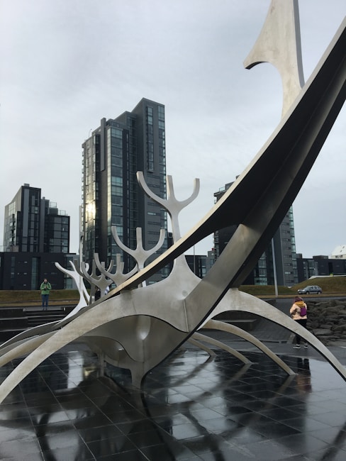 A modern art sculpture made of metal with sweeping lines and curved forms is set against the backdrop of tall, contemporary glass and steel buildings. The sculpture seems to have an abstract design reminiscent of a ship or creature. The ground is wet, suggesting recent rain, and there are a few people walking nearby, giving scale to the sculpture.