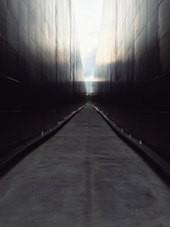 A long, narrow pathway is flanked by tall, dark walls that create a symmetrical, tunnel-like effect. The surface of the path is smooth, and small lights line the edges, leading the eye towards the vanishing point in the distance.