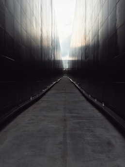 A long, narrow pathway is flanked by tall, dark walls that create a symmetrical, tunnel-like effect. The surface of the path is smooth, and small lights line the edges, leading the eye towards the vanishing point in the distance.