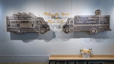 Black chalkboard drawings depict two food trucks with the words 'The Grilled Cheeserie' on a light blue wall. Above the trucks is a yellow cursive text that reads 'Melting the Hearts of Music City.' Below the wall, a wooden table with black chairs is adorned with a small vase of flowers.