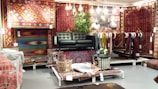A display area featuring a collection of intricately patterned rugs in various colors and designs. A dark green leather sofa is placed at the center with several hanging light bulbs above it, providing a cozy atmosphere. There are leafy green plants adding a natural touch, and an assortment of small decor items are arranged on the rug-covered platforms.