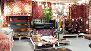A display area featuring a collection of intricately patterned rugs in various colors and designs. A dark green leather sofa is placed at the center with several hanging light bulbs above it, providing a cozy atmosphere. There are leafy green plants adding a natural touch, and an assortment of small decor items are arranged on the rug-covered platforms.