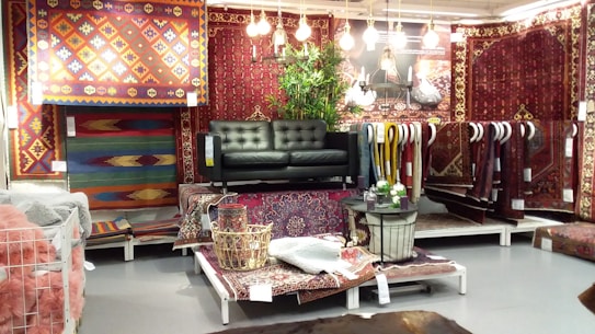 A display area featuring a collection of intricately patterned rugs in various colors and designs. A dark green leather sofa is placed at the center with several hanging light bulbs above it, providing a cozy atmosphere. There are leafy green plants adding a natural touch, and an assortment of small decor items are arranged on the rug-covered platforms.