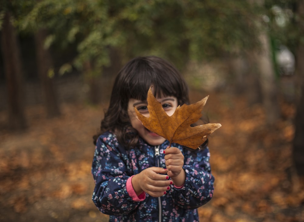 shallow focus photo of girl in blue and pink floral full-zip jacket holding maple leaf