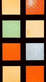 A grid of nine colorful glass panes, each with an ornate, embossed pattern. The glass panes vary in color, alternating between shades of orange, green, and blue.