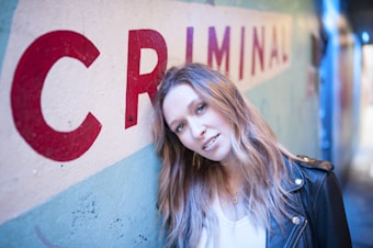 A person stands in a narrow hallway, leaning against a wall with the word 'CRIMINAL' painted in large red letters. The individual is wearing a black leather jacket and has long hair. The setting has a cool, urban vibe with blue and teal tones.