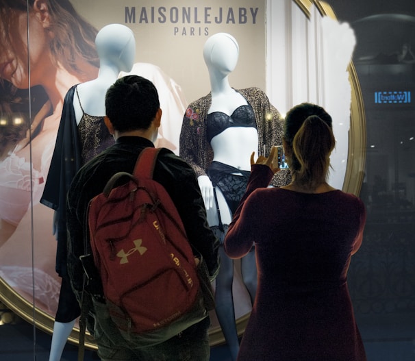 Two people stand in front of a store window display, observing mannequins dressed in lingerie. The storefront is branded 'MAISONLEJABY Paris'. One individual is taking a photo with a smartphone, while both focus on the display. The background features a large poster of a model.