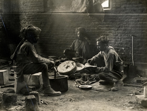Three individuals are engaged in a communal cooking activity inside a rustic, brick-walled room. The lighting is dim, hinting at a historical or vintage setting, with dust and light beams visible. The people appear to be preparing food over makeshift cooking implements, surrounded by various kitchen tools and equipment.