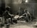 Three individuals are engaged in a communal cooking activity inside a rustic, brick-walled room. The lighting is dim, hinting at a historical or vintage setting, with dust and light beams visible. The people appear to be preparing food over makeshift cooking implements, surrounded by various kitchen tools and equipment.