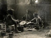 Three individuals are engaged in a communal cooking activity inside a rustic, brick-walled room. The lighting is dim, hinting at a historical or vintage setting, with dust and light beams visible. The people appear to be preparing food over makeshift cooking implements, surrounded by various kitchen tools and equipment.
