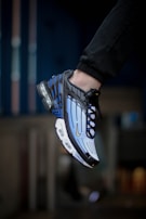 A close-up view of a sneaker being worn on a person's foot. The shoe has a striking design with a combination of blue, black, and white colors, featuring a visible air cushioning in the heel and distinct lines across the upper part. The background appears blurred, emphasizing the shoe.