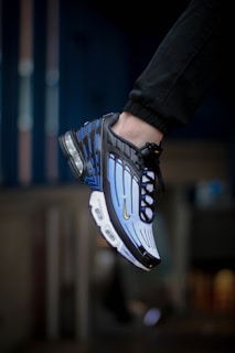 A close-up view of a sneaker being worn on a person's foot. The shoe has a striking design with a combination of blue, black, and white colors, featuring a visible air cushioning in the heel and distinct lines across the upper part. The background appears blurred, emphasizing the shoe.