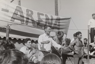 A group of men, some shirtless, are gathered around a platform or stage. A man in a white shirt appears to be speaking or gesturing to a crowd. A large sign with the word 'ARENA' is prominently displayed in the background. The scene seems to be set outdoors on a sunny day.