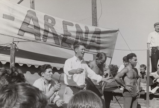 A group of men, some shirtless, are gathered around a platform or stage. A man in a white shirt appears to be speaking or gesturing to a crowd. A large sign with the word 'ARENA' is prominently displayed in the background. The scene seems to be set outdoors on a sunny day.