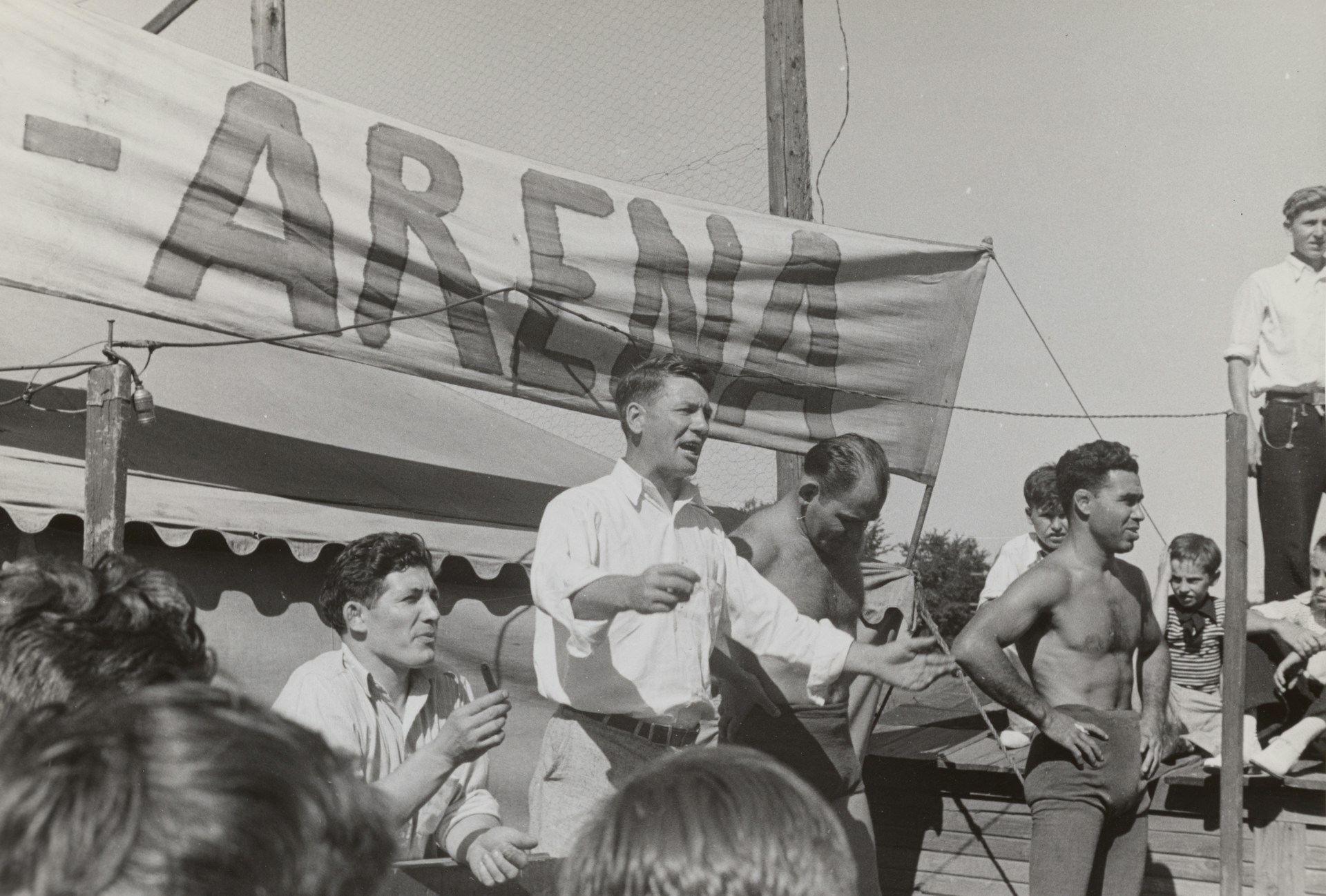 A group of men, some shirtless, are gathered around a platform or stage. A man in a white shirt appears to be speaking or gesturing to a crowd. A large sign with the word 'ARENA' is prominently displayed in the background. The scene seems to be set outdoors on a sunny day.