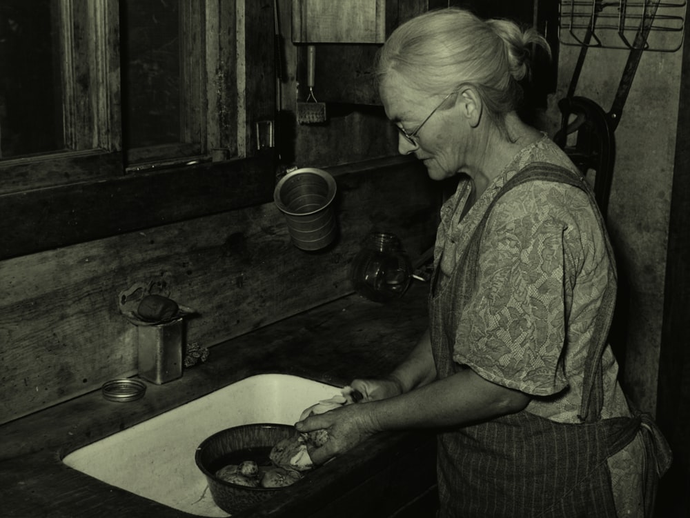 grayscale photography of woman standing beside sink