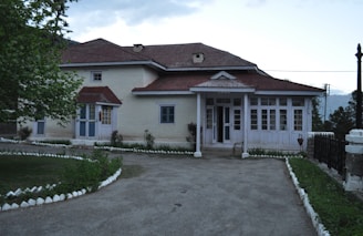 An old, rustic house with a red roof and white walls, surrounded by a small, neatly maintained garden. The house has multiple windows and a couple of small porches, with a paved walkway leading to the entrance. The surrounding area is lush with greenery and trees.