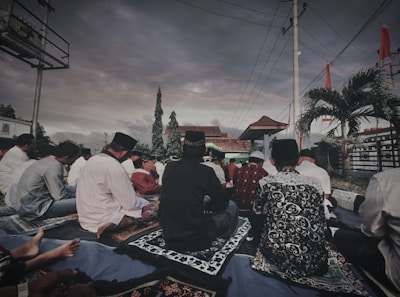 A group of people is seated outdoors on prayer mats under a cloudy sky. The setting includes trees, a tiled roof structure, and palm trees. The participants appear to be engaged in a communal activity, some wearing traditional attire and head coverings.