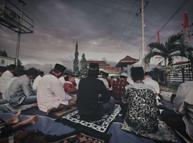 A group of people is seated outdoors on prayer mats under a cloudy sky. The setting includes trees, a tiled roof structure, and palm trees. The participants appear to be engaged in a communal activity, some wearing traditional attire and head coverings.