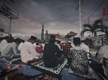 A group of people is seated outdoors on prayer mats under a cloudy sky. The setting includes trees, a tiled roof structure, and palm trees. The participants appear to be engaged in a communal activity, some wearing traditional attire and head coverings.