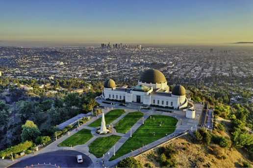 A large observatory with distinct domes stands on a hill surrounded by lush greenery, overlooking a sprawling cityscape. The skyline in the distance features numerous skyscrapers under a clear blue sky as the sun casts a warm glow over the scene.