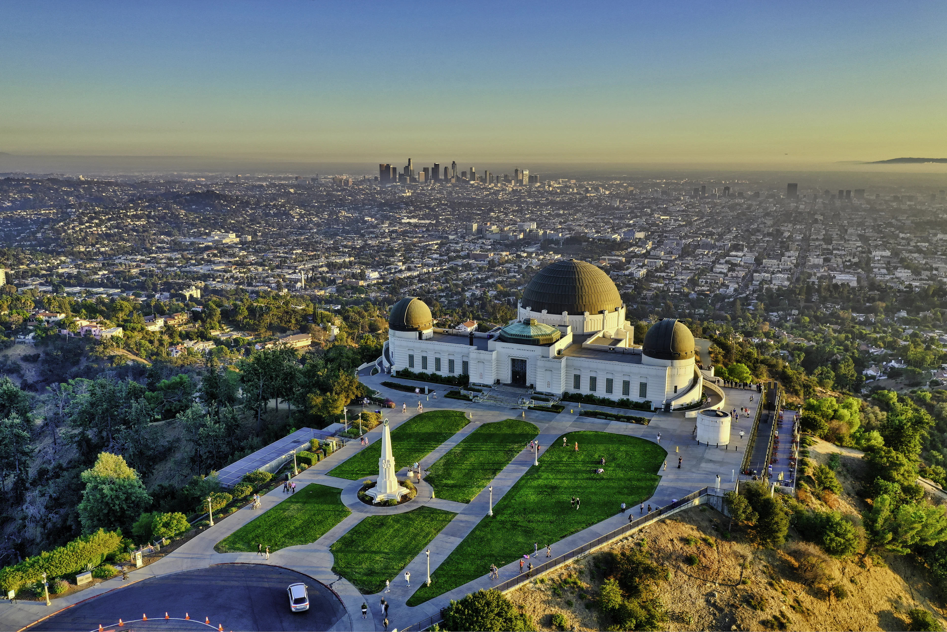 Griffith Observatory and its copper domes seen from above at golden hour, the lawns and paths of Griffith Park below and the Downtown Los Angeles skyline rising out of the haze on the horizon.