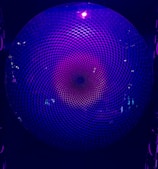 A circular geometric pattern with a repetitive, hypnotic design illuminated by purplish-blue light. The pattern creates a sense of depth and movement, resembling a vortex or spiral.