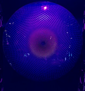 A circular geometric pattern with a repetitive, hypnotic design illuminated by purplish-blue light. The pattern creates a sense of depth and movement, resembling a vortex or spiral.
