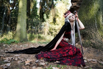 A person with long white hair, wearing a red and black gown with a black cape, is kneeling beside a large tree in a forest. They are wearing a red crown and holding a long sword upright. The ground is covered with dry leaves, and the background is filled with tall trees and greenery.