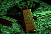 A packet of Zig-Zag branded rolling papers with a vintage illustration on the front is surrounded by other similar products on a textured surface. The lighting casts a greenish hue over the scene, creating a moody atmosphere.