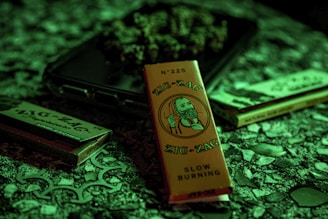 A packet of Zig-Zag branded rolling papers with a vintage illustration on the front is surrounded by other similar products on a textured surface. The lighting casts a greenish hue over the scene, creating a moody atmosphere.